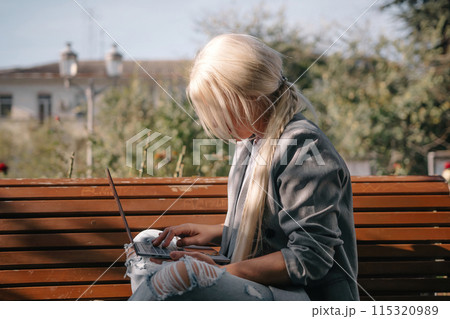 A blonde woman sits on a bench with a laptop open in front of her. She is wearing a gray jacket and has her hair in a ponytail. The scene suggests a casual and relaxed atmosphere. A blonde woman sits on a bench with a laptop open in front of her. She is wearing a gray jacket and has her hair in a ponytail. The scene suggests a casual and relaxed atmosphere. 115320989