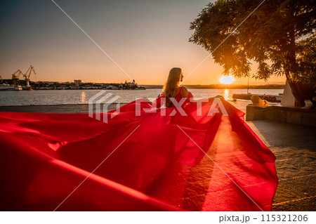 Sunrise red dress. A woman in a long red dress against the backdrop of sunrise, bright golden light of the sun's rays. The concept of femininity, harmony. 115321206