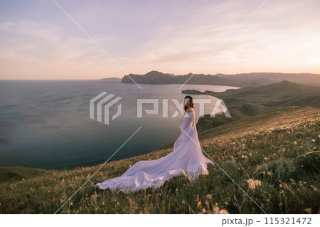 A woman in a white dress stands on a grassy hill overlooking the ocean 115321472