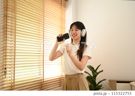 Happy young asian woman having fun listening to music and singing in living room Happy young asian woman having fun listening to music and singing in living room 115322755