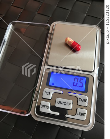 Precise stylish metal pocket scale with blue display. The red medical capsule is on the weight measurement. Close-up 115323120