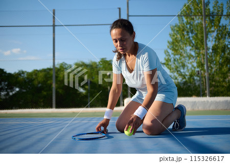 Mature woman playing tennis on outdoor court kneeling to pick up tennis ball 115326617