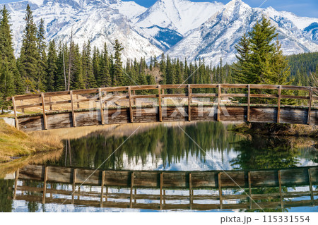 Wooden arch footbridge in Cascade Ponds park in autumn sunny day, snow-covered Mount Astley reflection on the water surface. Banff National Park, Canadian Rockies, Alberta, Canada. 115331554