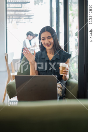 A woman is sitting in a chair with a laptop in front of her. She is smiling and waving at the camera. She is holding a cup of coffee 115342610
