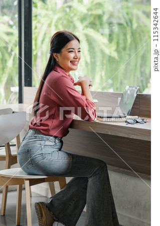 A woman in a pink shirt and blue jeans sits at a table with a laptop and a cup. She is smiling and she is enjoying her time 115342644