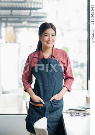 Smiling female barista in a denim apron standing in a modern cafe, ready to serve customers. Bright and airy coffee shop interior. 115342645