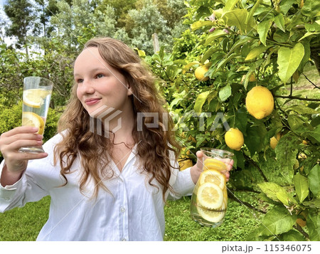 it's just a lemon paradise, a teenage girl stands against the backdrop of a lemon tree with lemons and drinks lemonade from a glass glass in her other hand, she holds a jug of drink it's just a lemon paradise, a teenage girl stands against the backdrop of a lemon tree with lemons and drinks lemonade from a glass glass in her other hand, she holds a jug of drink 115346075