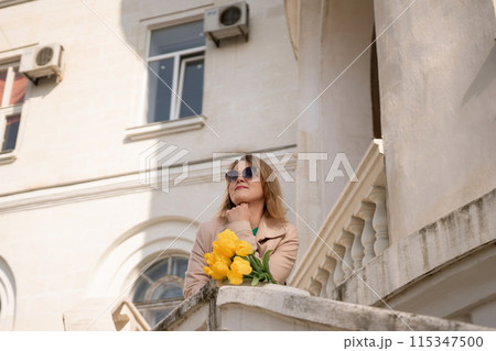A woman wearing sunglasses and holding a bouquet of yellow flowers stands on a balcony. The scene is peaceful and serene, with the woman looking out over the city. 115347500