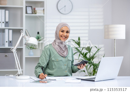 Smiling businesswoman in hijab working on laptop and holding a calculator in modern office setting. Smiling businesswoman in hijab working on laptop and holding a calculator in modern office setting. 115353517
