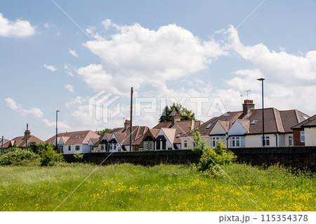 白い雲の浮かぶ明るい日差しの下の三角屋根の家並みと黄色の花が咲くフィールド ロンドン郊外にて 白い雲の浮かぶ明るい日差しの下の三角屋根の家並みと黄色の花が咲くフィールド ロンドン郊外にて 115354378