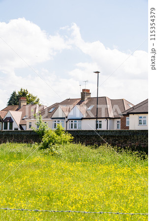 白い雲の浮かぶ明るい日差しの下の三角屋根の家並みと黄色の花が咲くフィールド ロンドン郊外にて 白い雲の浮かぶ明るい日差しの下の三角屋根の家並みと黄色の花が咲くフィールド ロンドン郊外にて 115354379