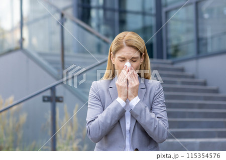 Businesswoman sneezing outside office building due to seasonal allergies. Professional woman suffering from cold or flu symptoms 115354576