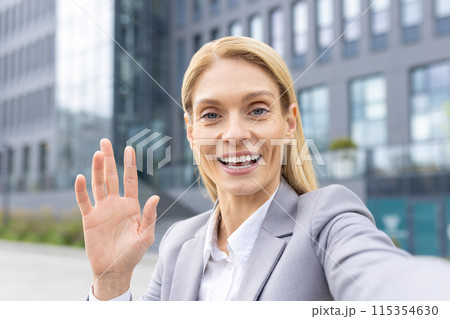 A cheerful businesswoman in a suit waving and smiling at the camera in front of a contemporary office building. 115354630