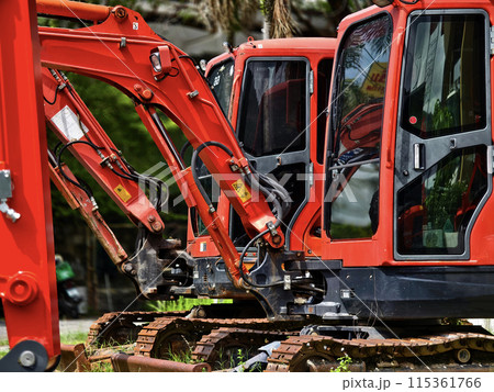 Red large excavators are stationed in a green field in front of a building 115361766