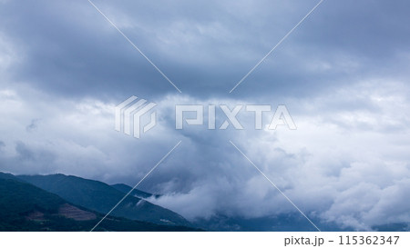 雨の日の山頂を、覆いかぶさりながら流れていく雨雲 115362347