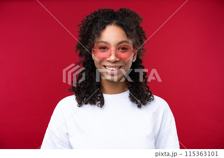 Portrait of a young girl smiling at the camera while standing against a red background. Happy black woman embracing her natural hair. 115363101