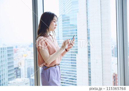 オフィスの窓辺でスマホを見る女性 オフィスの窓辺でスマホを見る女性 115365768