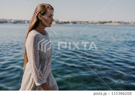 A woman is walking on the beach wearing a dress. The water is calm and the sky is clear. 115366208