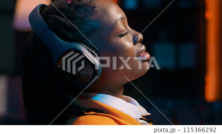 College student sitting laid back after working on class project, relaxing with ambience music on headphones at home desk. African american girl chilling with modern tunes on headset. Camera A. College student sitting laid back after working on class project, relaxing with ambience music on headphones at home desk. African american girl chilling with modern tunes on headset. Camera A. 115366292