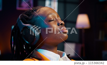 Carefree student finishes creating a presentation and relaxes with music on audio headset, resting after long researching session. African american girl chilling with ambience tunes. Camera B. 115366342