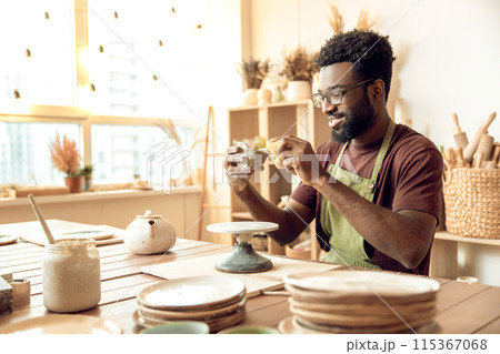 Man in apron working in the pottery studio and looking involved Man in apron working in the pottery studio and looking involved 115367068