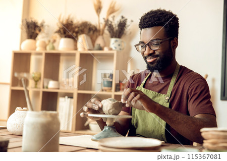 Dark-skinned man in eyeglasses shaping mug from clay and looking focused 115367081