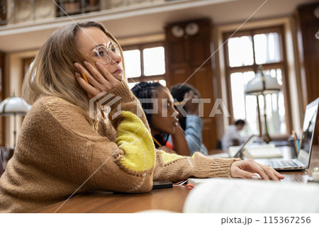 Blonde young woman sitting at the table in the library and feeling tired Blonde young woman sitting at the table in the library and feeling tired 115367256