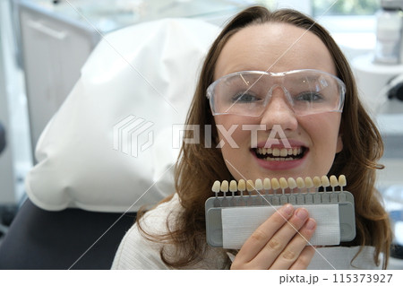 Smiling young woman. Cosmetological teeth whitening in a dental clinic. selection of the tone of the implant tooth Smiling young woman. Cosmetological teeth whitening in a dental clinic. selection of the tone of the implant tooth 115373927