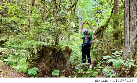 a girl in a Green T-shirt stands with her back in the jungle raised her hands up she shows her fingers the heart of a teenagers travel in Canada on Vancouver Island in a forest covered with moss ferns a girl in a Green T-shirt stands with her back in the jungle raised her hands up she shows her fingers the heart of a teenagers travel in Canada on Vancouver Island in a forest covered with moss ferns 115374117