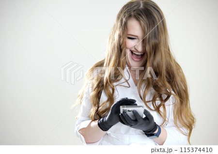 happy contented cheerful woman doctor workwear white laughs open mouth looks into mobile phone learns news latest technology communication online conference meeting with friends and gorgeous hair 115374410