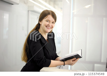 Image of young woman, company worker smiling and holding notepad standing against Image of young woman, company worker smiling and holding notepad standing against white background 115374549