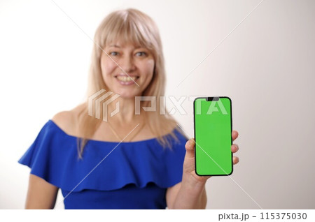 supply demand An adult woman 45-50 with blond hair in a blue dress is standing with mobile phone Green screen chroma key smiles advertising any products Sincere smile beautiful face blurred background supply demand An adult woman 45-50 with blond hair in a blue dress is standing with mobile phone Green screen chroma key smiles advertising any products Sincere smile beautiful face blurred background 115375030