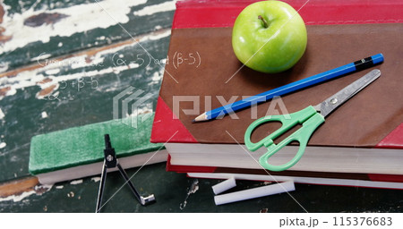 A green apple sits atop a stack of books next to a blue pencil, compass, and scissors 115376683