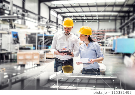 Two project managers standing in modern industrial factory. Manufacturing facility with robotics, robotic arms and automation. Storing products and materials in warehouse. 115381493