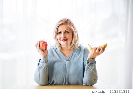 Portrait of beautiful young overweight woman at home, choosing healthy food, eating fruits. 115381531