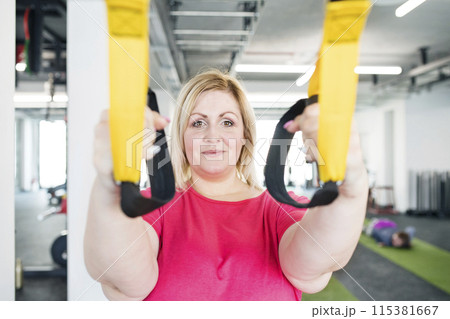 Overweight woman exercising in gym, doing suspension training, total resistance exercise. Personal trainer couching her and helping her. Overweight woman exercising in gym, doing suspension training, total resistance exercise. Personal trainer couching her and helping her. 115381667