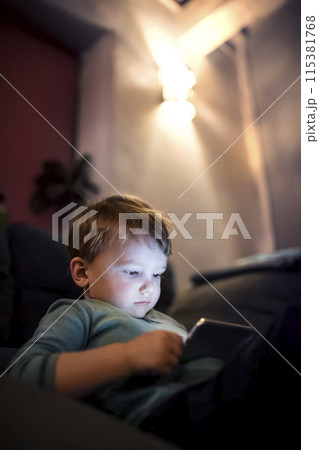 Cute boy sitting on bed watching something on smartphone. Children's screen time. 115381768