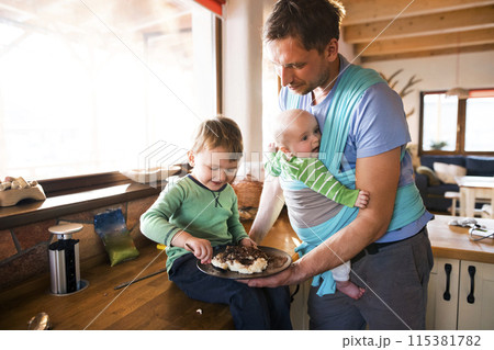 Father holding small baby in baby sling. Older brother eating porridge on kitchen counter. Unconditional parental love, family moment. 115381782