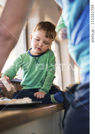 Father holding small baby in baby sling. Older brother eating porridge on kitchen counter. Unconditional parental love, family moment. 115381784