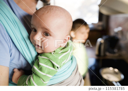 Father holding small baby in baby sling. Older brother stirring porridge in pot. Unconditional parental love, family moment. Father holding small baby in baby sling. Older brother stirring porridge in pot. Unconditional parental love, family moment. 115381785
