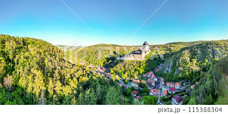Panoramic view of Karlstejn Castle surrounded by lush greenery and a quaint village under a clear blue sky during a summer morning. 115388304