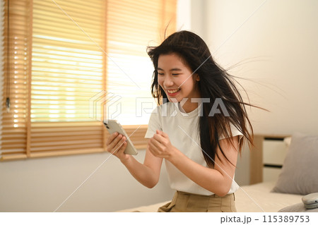 Young Asian woman reading text message on her phone with happy pleased face expression Young Asian woman reading text message on her phone with happy pleased face expression 115389753