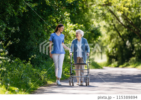 Female caregiver and senior woman with walker on walk in nature. Nurse and elderly woman enjoying a warm day in nursing home, public park. Female caregiver and senior woman with walker on walk in nature. Nurse and elderly woman enjoying a warm day in nursing home, public park. 115389894