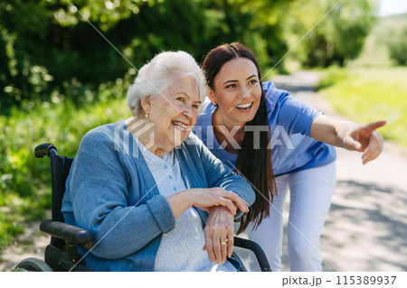 Female caregiver pushing senior woman in wheelchair. Nurse and elderly woman enjoying a warm day in nursing home, public park. 115389937