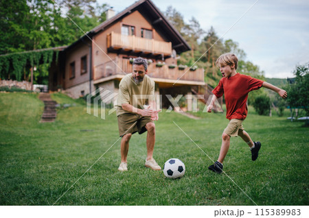Dad having fun with young son, playing football, running and kicking the ball. Playing on a lawn in front of their house. Fatherhood and Father's Day. Dad having fun with young son, playing football, running and kicking the ball. Playing on a lawn in front of their house. Fatherhood and Father's Day. 115389983