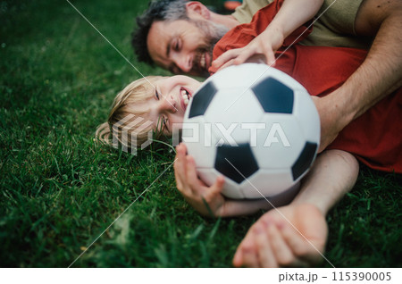 Dad lying on grass with son, laughing and having fun. Father and boy playing football, catching each other, falling to the ground. Fatherhood and Father's Day. Dad lying on grass with son, laughing and having fun. Father and boy playing football, catching each other, falling to the ground. Fatherhood and Father's Day. 115390005