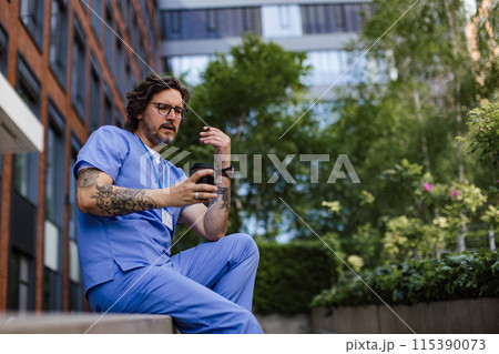 Healthcare worker taking a break, smoking a cigarette outdoors and scrolling on phone, in front of the hospital building. 115390073