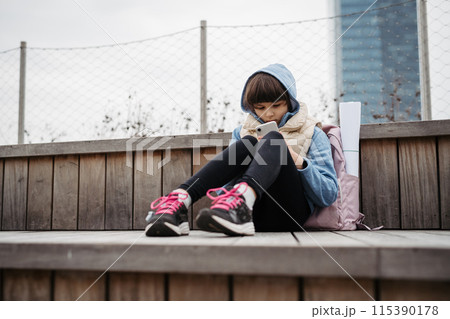 Sad girl phone calling, sitting in front of school building, waiting for parents. Young schoolgirl with no friends, feeling lonely. 115390178