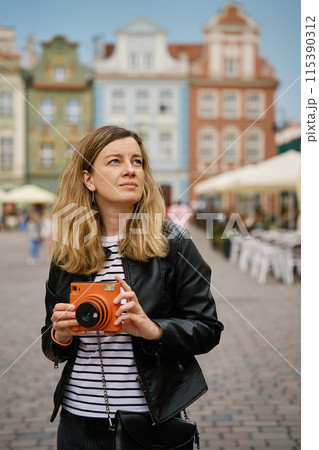 Woman taking picture using orange instant camera. Tourist captures memories during travel with vintage camera. Female walking around city street 115390312