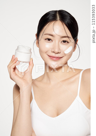 an Asian woman with cream on her face and holding cosmetics 115393033